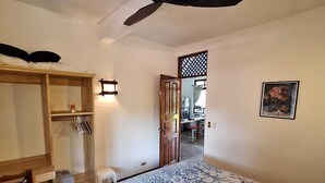 Quarto com cama, escrivaninha de madeira, cadeira, guarda-roupa, ventoinha no teto e porta que dá para outro cômodo.