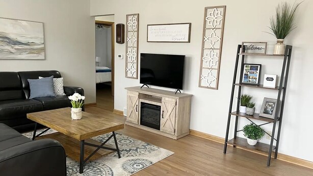 Un salon avec un canapé en cuir noir, une table basse en bois, un téléviseur à écran plat, une cheminée et une étagère décorative agrémentée de plantes et de tableaux.