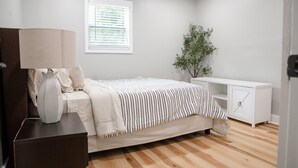 Un dormitorio con cama, mesita de noche, lámpara, ventana y una planta.