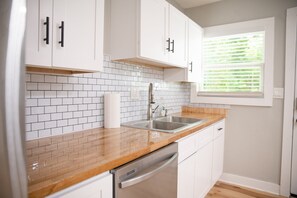 Une cuisine avec des meubles blancs, un plan de travail en bois, un évier en acier inoxydable et une fenêtre avec des stores.