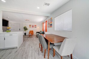 A modern kitchen with white cabinetry, a dining area with a wooden table and grey upholstered chairs, and a living area with a sofa and orange armchair.