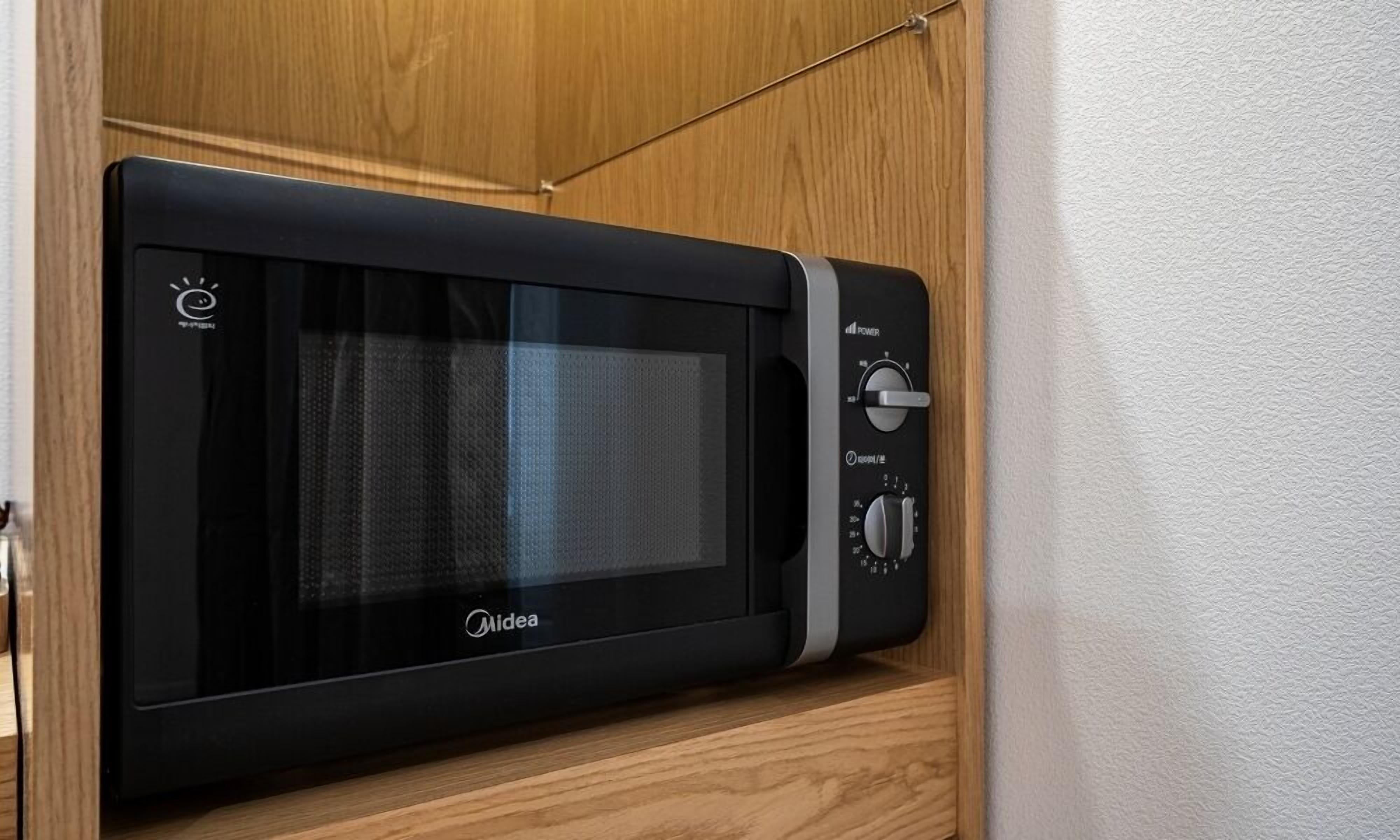 Un four à micro-ondes Midea noir, doté d’un écran numérique et de boutons de contrôle de la puissance, est posé sur une étagère en bois dans une cuisine.