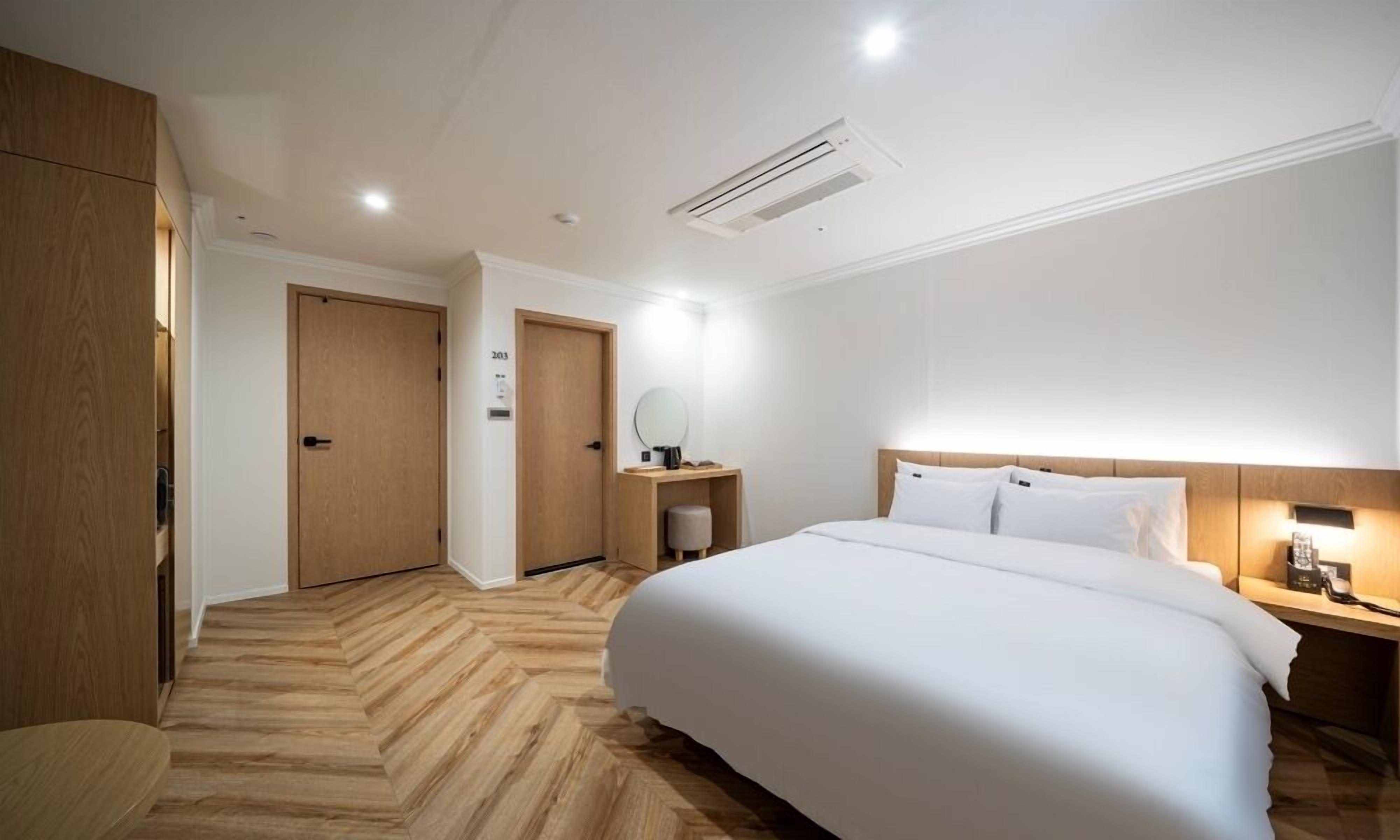 A modern hotel room with a large bed, a bedside table with a lamp, a small stool, and a wooden wardrobe.