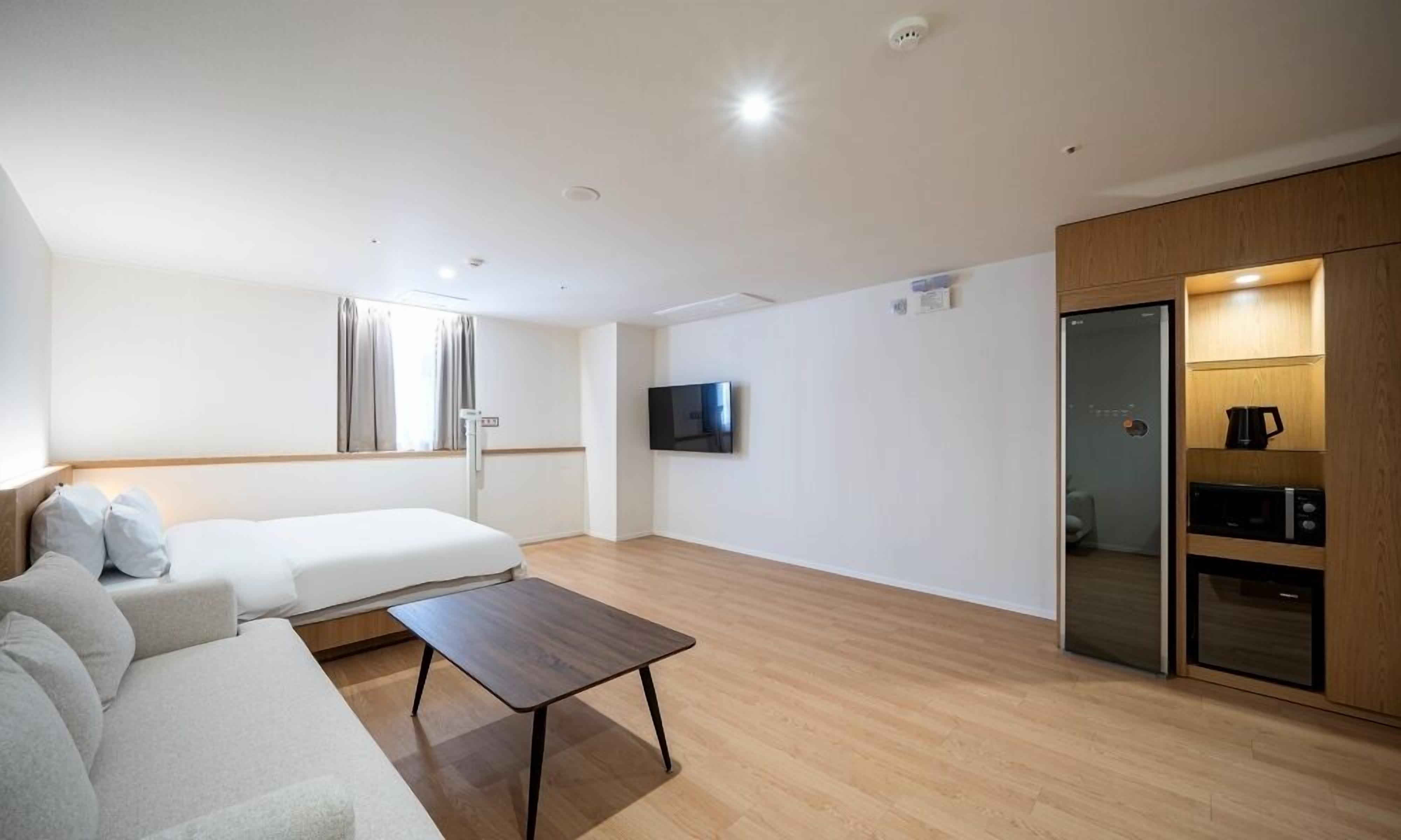 A modern hotel room with a bed, sofa, TV, wooden floor, and built-in kitchen.