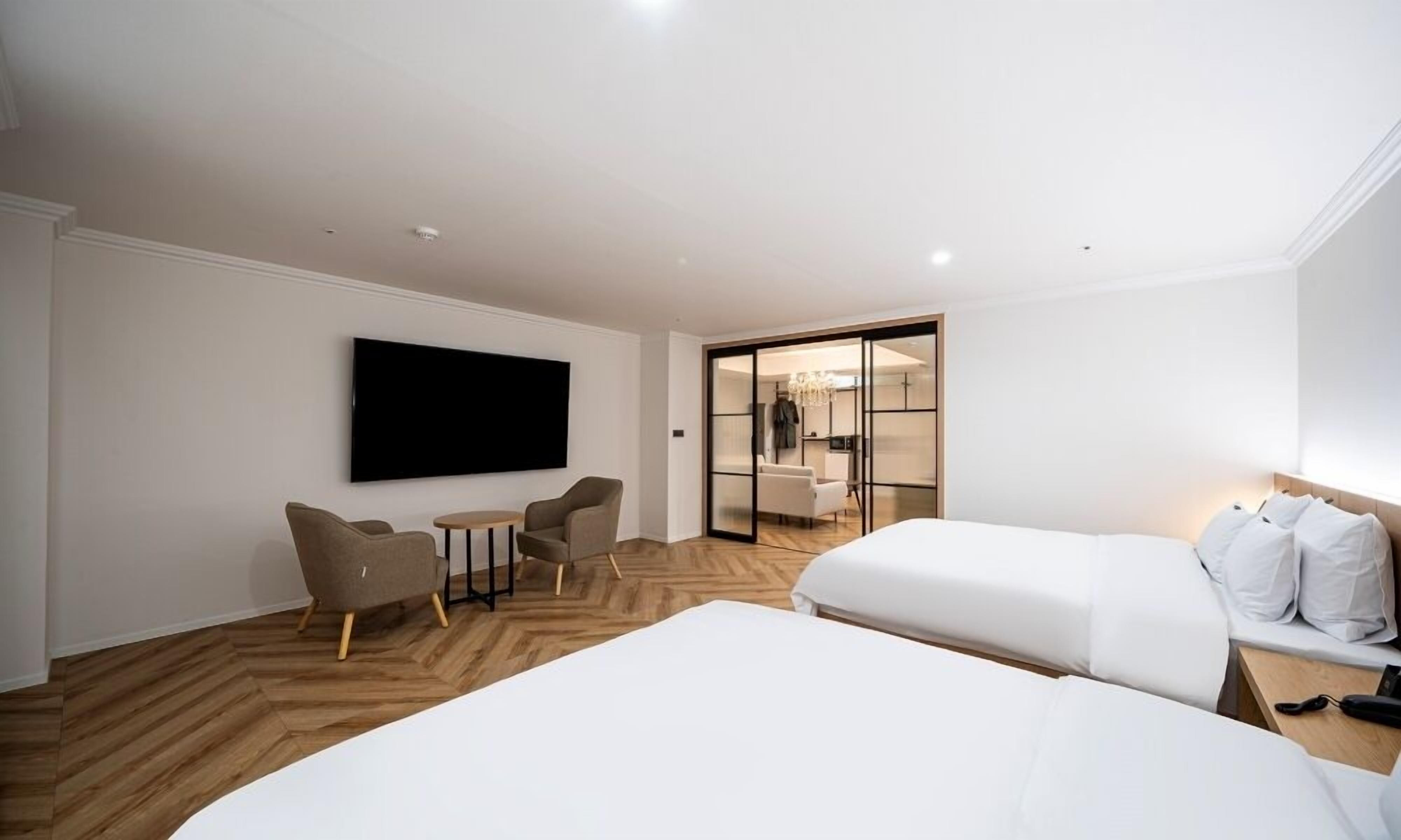 A hotel room with a large bed, a TV mounted on the wall, a seating area with chairs and a table, and a sliding door leading to a bathroom.