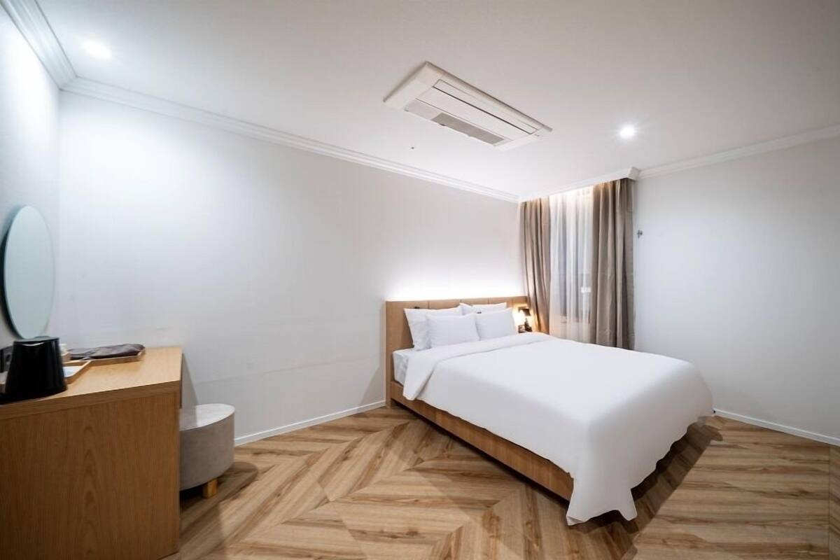 A modern hotel room with a wooden herringbone floor, a large bed with white linens, a wooden nightstand, a round mirror, and a ceiling light fixture.
