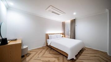 Habitación de hotel moderna con suelo de madera en patrón de espiga, una cama grande con ropa blanca, una mesita de noche de madera, un espejo redondo y una lámpara de techo.