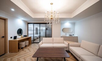 A modern living room with a wooden coffee table, beige sofas, a chandelier, and a sliding glass door leading to a bedroom.
