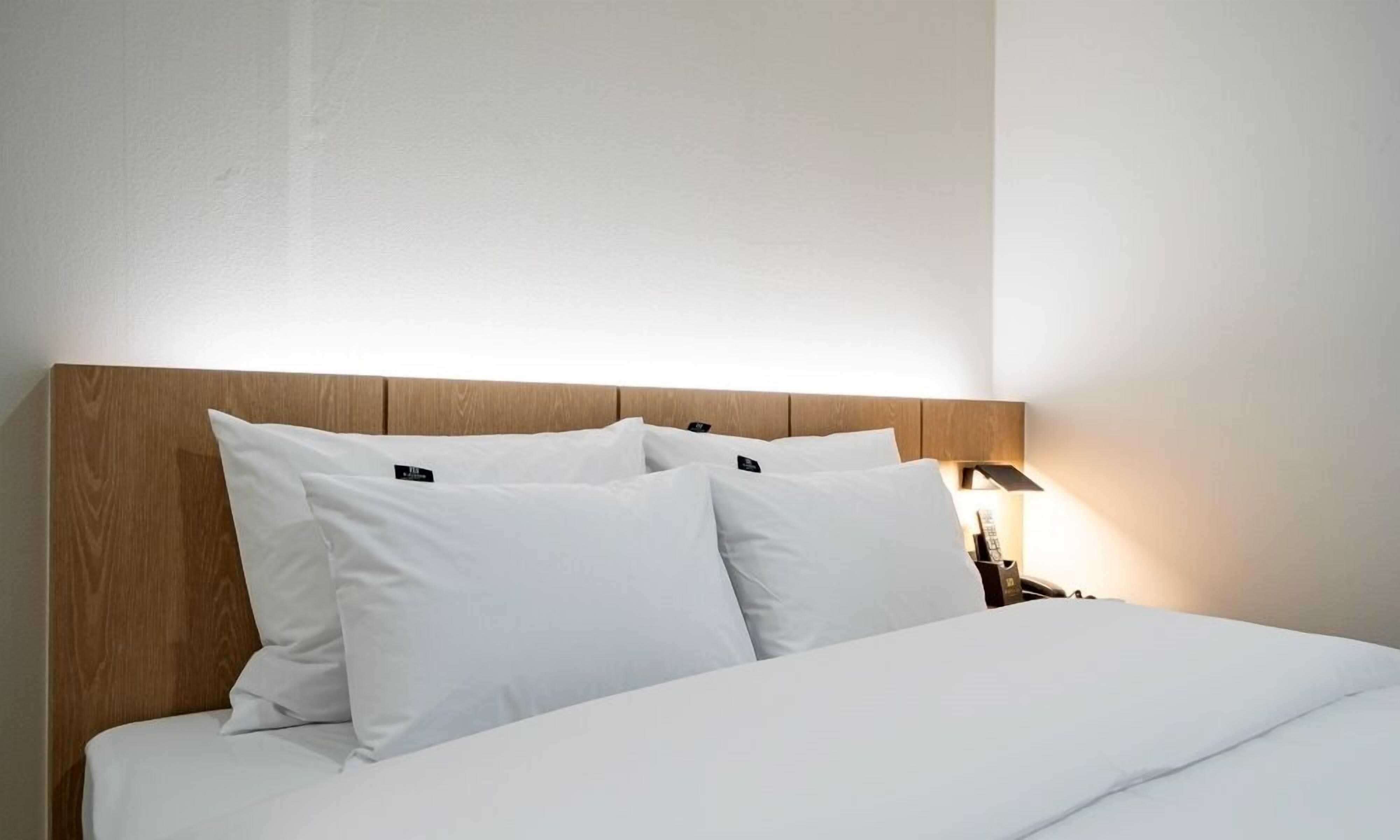 Uma cama bem arrumada com roupa de cama e almofadas brancas, uma cabeceira de madeira e um abajur.