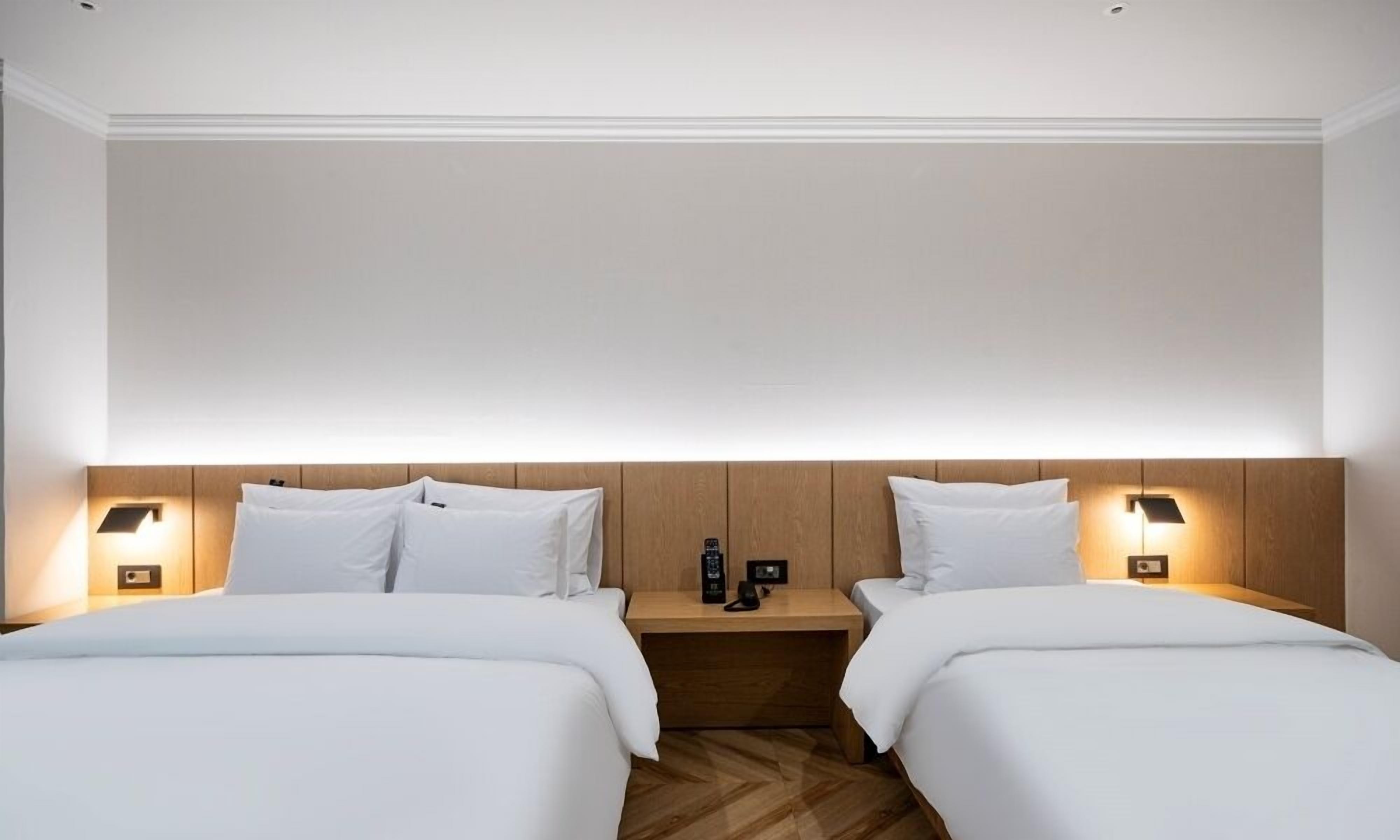 A hotel room with two beds, wooden headboards, bedside lamps, and a nightstand with a bottle and a phone.