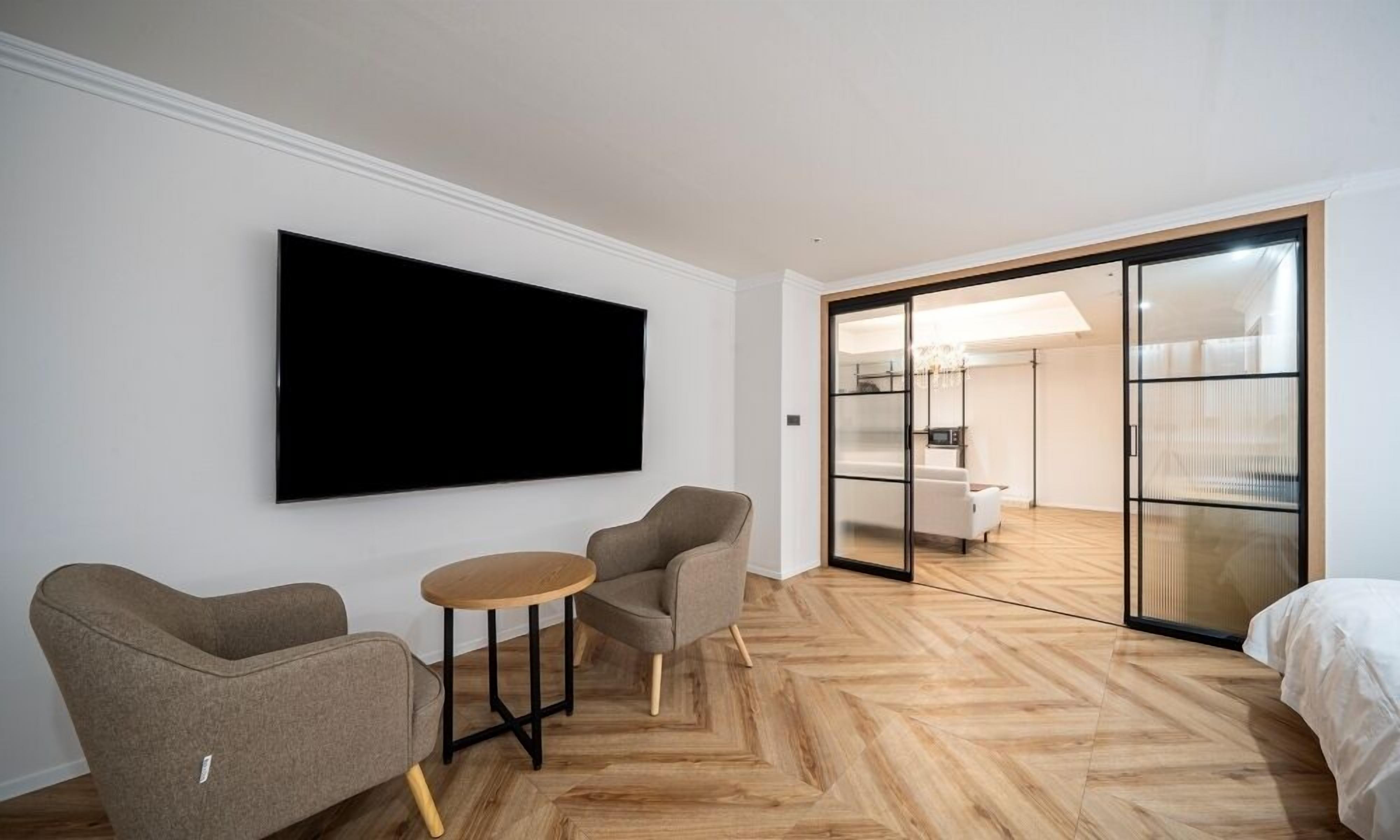 A modern hotel room with a large flat-screen TV, two armchairs, a small wooden table, and a sliding glass door leading to a bedroom.