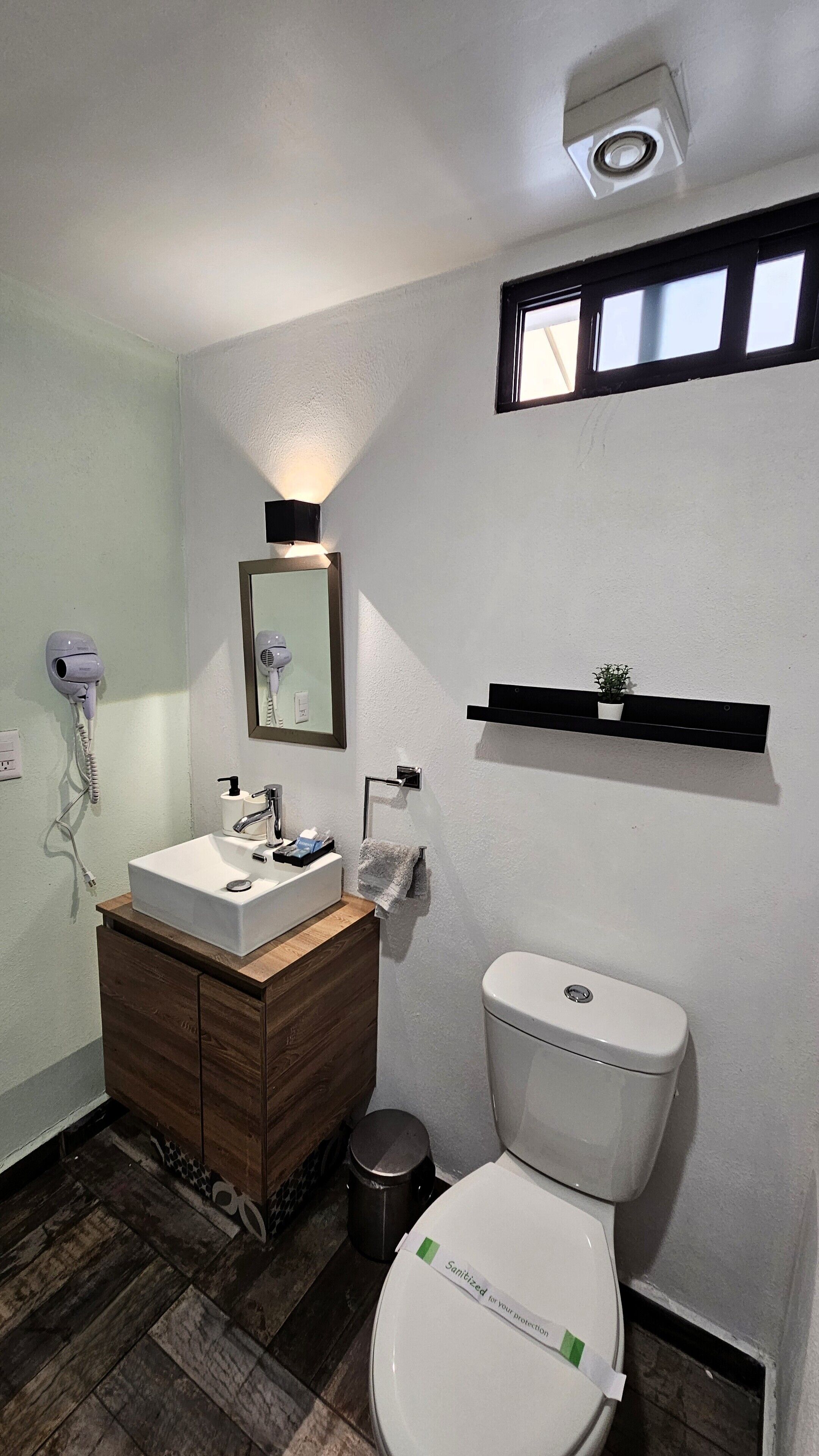A bathroom with a white sink, mirror, and toilet. There is a hairdryer on the wall, a small trash can, and a shelf with a plant.