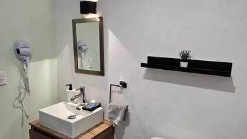 A bathroom with a white sink, mirror, and toilet. There is a hairdryer on the wall, a small trash can, and a shelf with a plant.