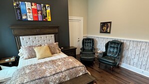Een slaapkamer met een knopen hoofdeinde, een groot bed met behang, twee leren fauteuils, een nachtkastje en een wandplank met boeken.