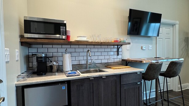 Een moderne keuken met een ingebouwde magnetron, koffiezetapparaat en spoelbak. Er is een televisie aan de muur bevestigd en twee barkrukken.