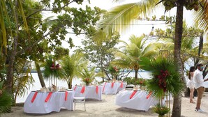 Une cérémonie de mariage en plein air, avec des tables blanches et des rubans rouges, entourée de palmiers et offrant une vue sur la plage.