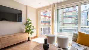 Un salon moderne doté d’un téléviseur à écran plat, de meubles en bois, d’une plante en pot et de grandes fenêtres offrant une vue sur les bâtiments voisins.
