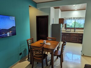 A dining area with a wooden table and chairs, a television displaying a starry night scene, and a kitchen with wooden cabinets and a window. - Romantic Private Villa w/ Hot Tub, Pool Access - Near La Fortuna and Arenal (La Fortuna)