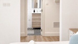 Un dormitorio con cama, mesita de noche, almohada azul, pared blanca, suelo de madera, baño con espejo, toalla y suelo de baldosas.