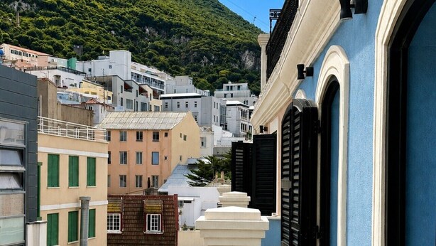 Una calle urbana estrecha con edificios, un edificio azul con contraventanas negras, y una colina al fondo.