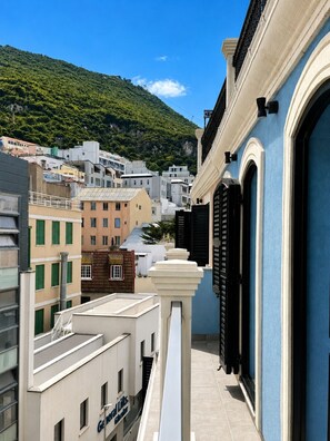 Uma rua urbana estreita com edifícios, incluindo um prédio azul com persianas pretas, e uma encosta ao fundo.