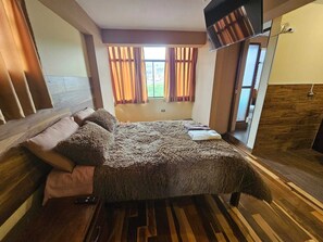 Un dormitorio con una cama grande, piso de madera, una ventana con cortinas y un televisor montado en la pared.