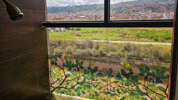 A bathroom with a large window overlooking a cityscape and a green area with trees. - beautiful and warm apartment,  5 minutes from Cusco airport (Cusco)