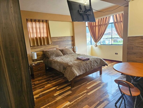 Un dormitorio con piso de madera, una cama con manta marrón, un televisor montado en la pared y un ventanal con cortinas.