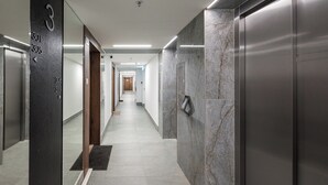 A modern hallway with a marble wall, a black pillar with room numbers, and an elevator. - Bright Crystal Premium Apartment (Warszawa)