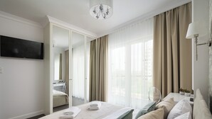 A modern hotel room with a large bed, a flat-screen TV, a desk with a lamp, and a view of the city through large windows. - Bright Crystal Premium Apartment (Warszawa)