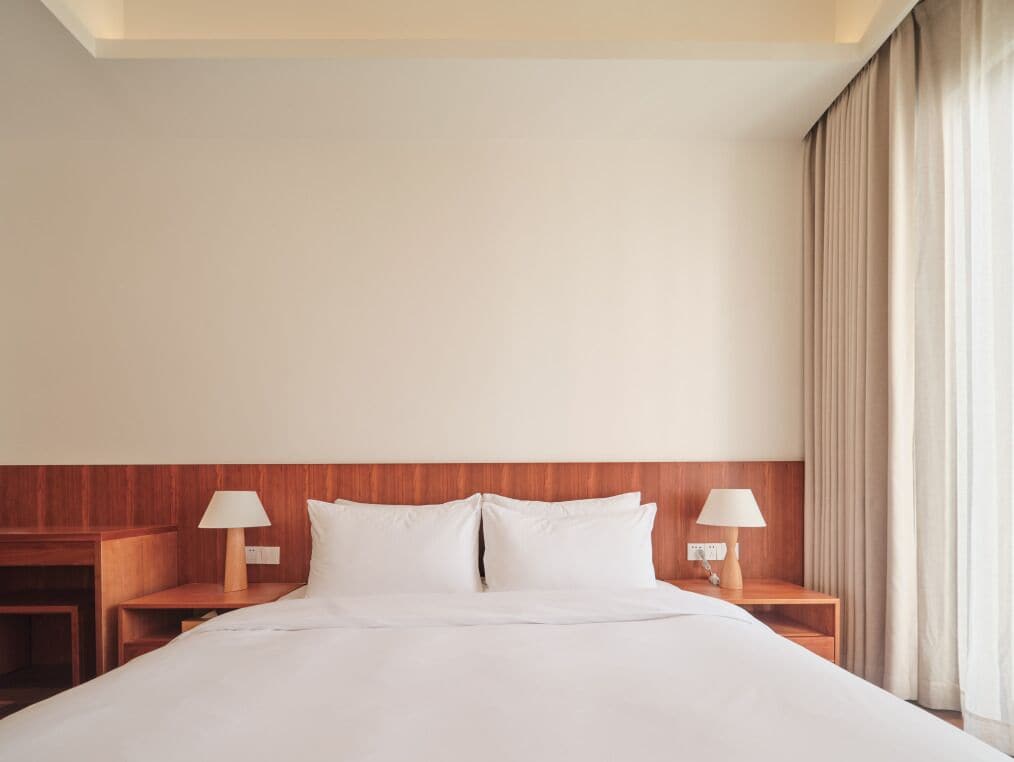 Habitación de hotel con una cama grande, cabecera de madera, dos mesitas de noche con lámparas y un ventanal con cortinas.