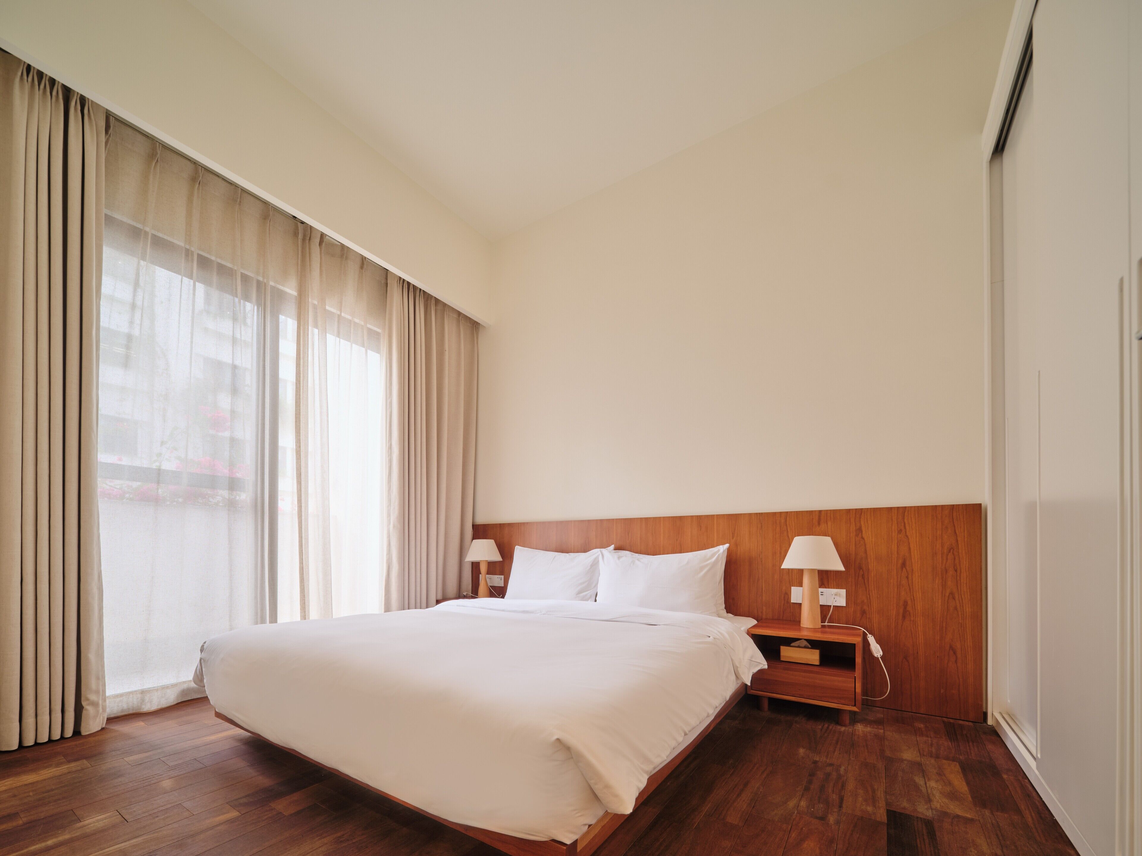 A hotel room with a large bed, wooden headboard, two bedside tables with lamps, and a large window with curtains.