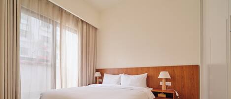 A hotel room with a large bed, wooden headboard, two bedside tables with lamps, and a large window with curtains.