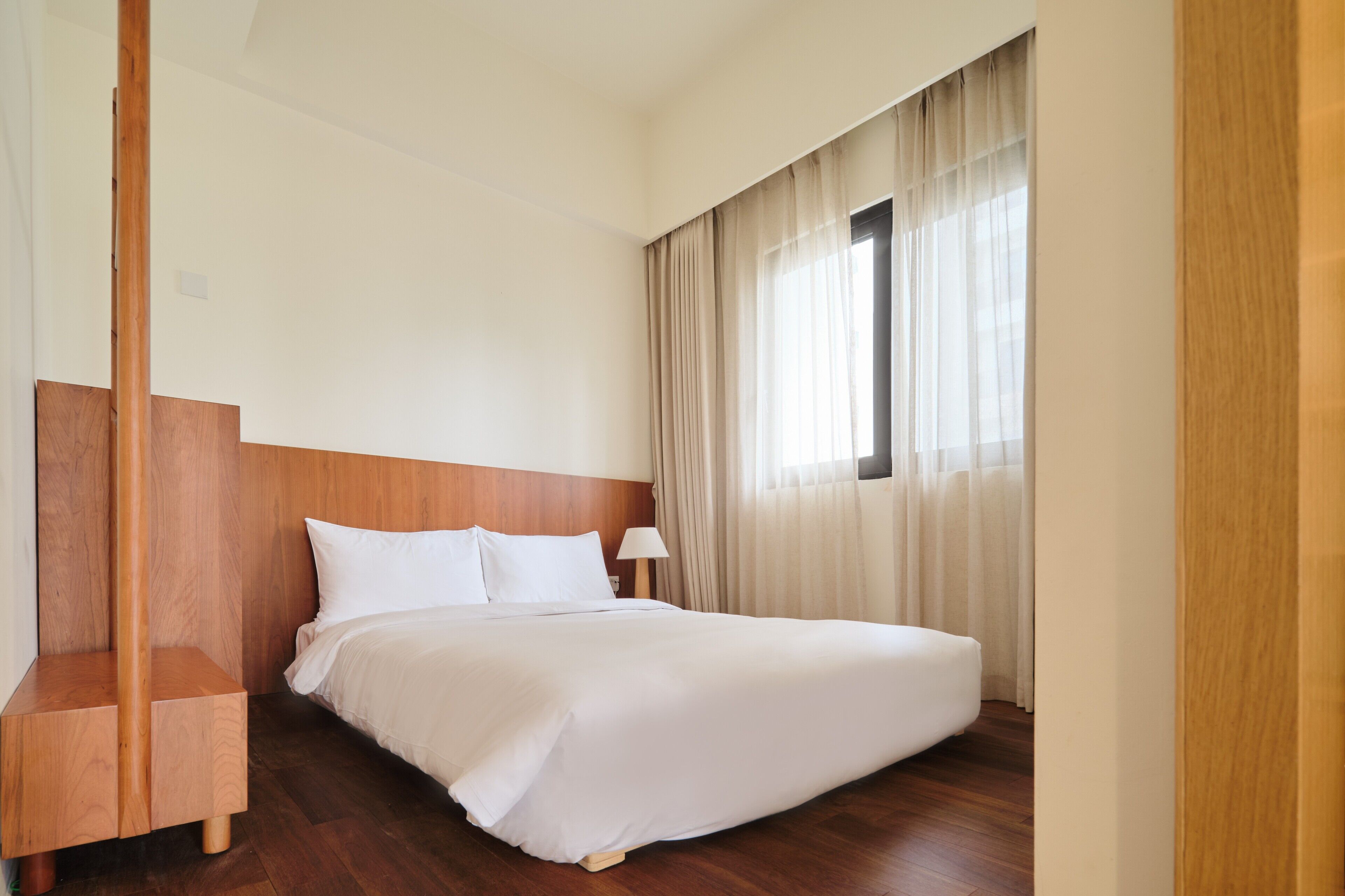 Una camera d'albergo con una testiera in legno, un letto grande con lenzuola bianche, un comodino, una lampada e una finestra con tende.