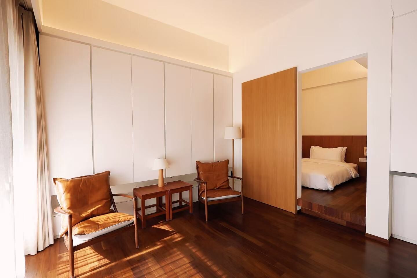 Een hotelkamer met een houten vloer, een bed met wit beddengoed, twee lederen fauteuils, een klein houten tafeltje met een lamp, en een groot raam met gordijnen.
