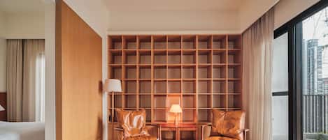 A modern hotel room with a wooden floor, a large window, a sofa, two chairs, and a small table with lamps.