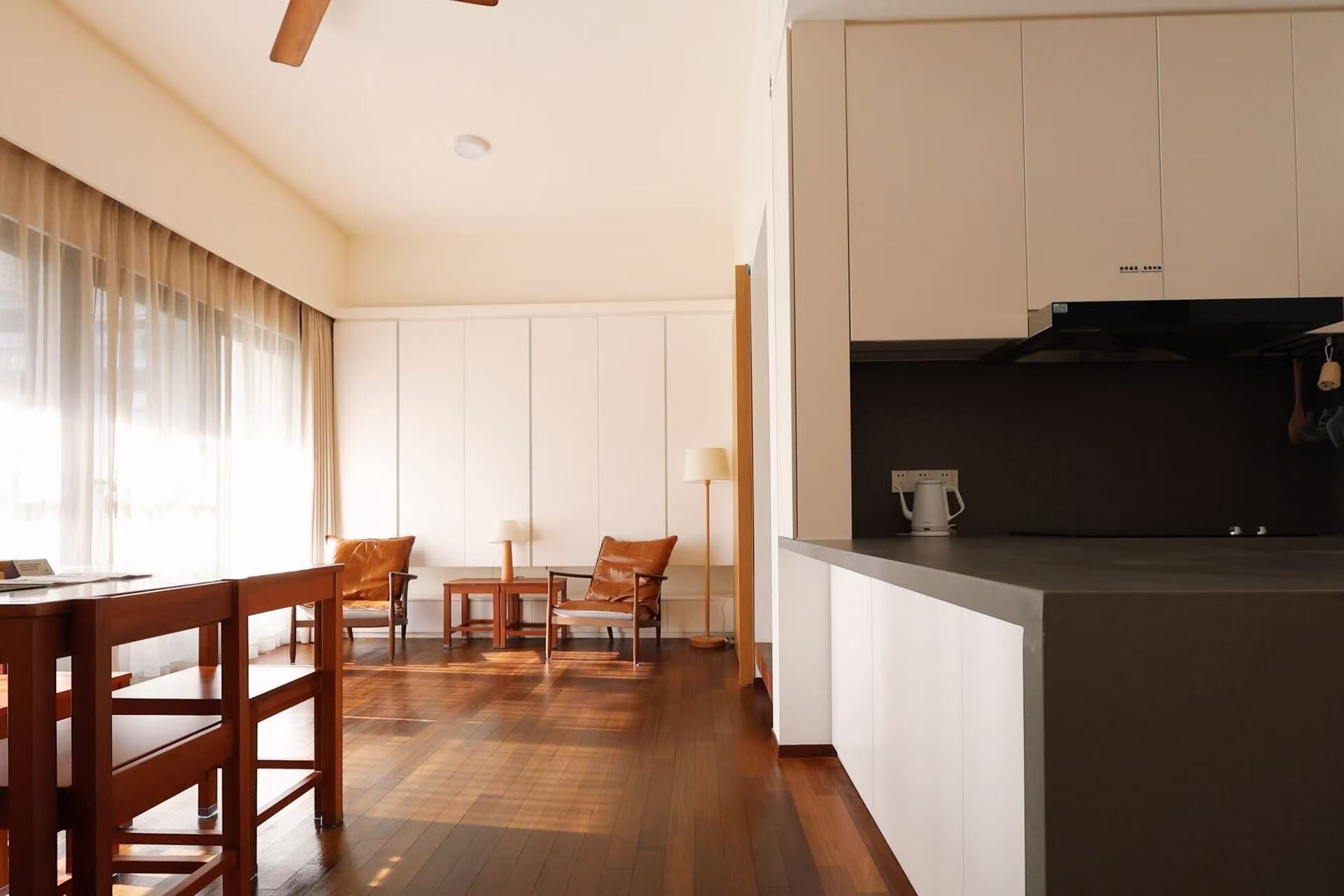 Una habitación de hotel moderna con una mesa de comedor de madera, sillas, una zona de cocina con cafetera y un amplio ventanal con cortinas translúcidas.