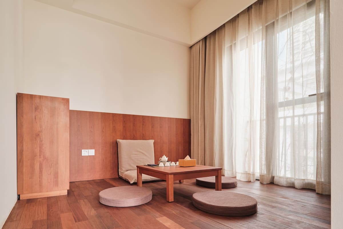 Ein Zimmer mit Holzboden, einem Holztisch mit Sitzkissen, einem Stuhl und einem Fenster mit durchsichtigen Vorhängen.