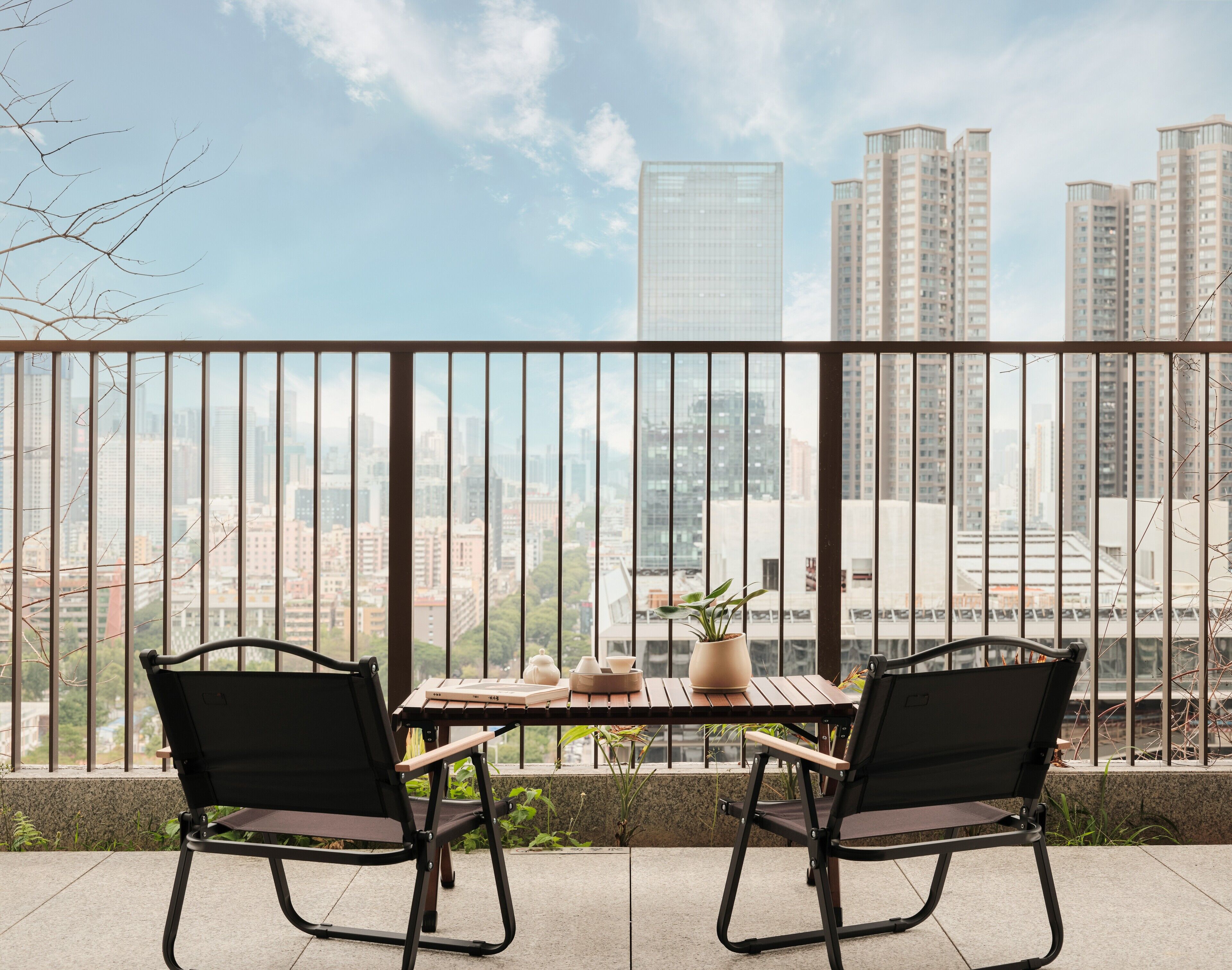 A balcony with two black chairs and a wooden table, overlooking a cityscape with tall buildings.