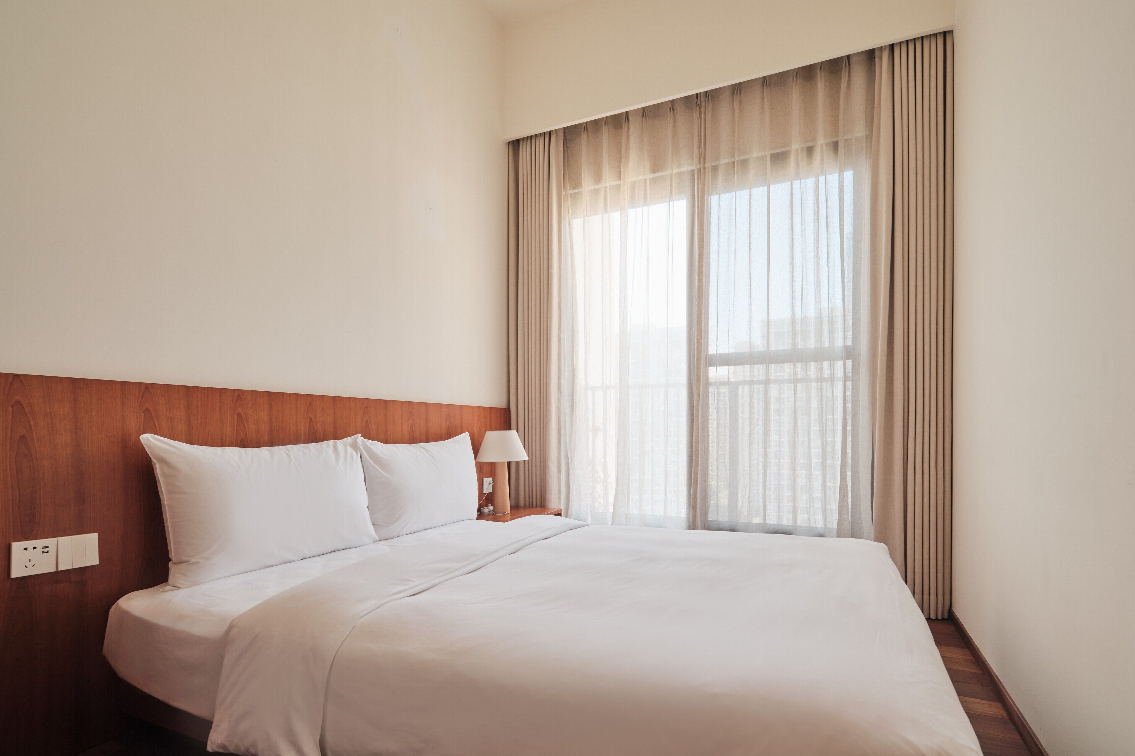 Una cama bien hecha con sábanas blancas, un cabecero de madera, una mesita de noche con una lámpara y una ventana con cortinas traslúcidas.