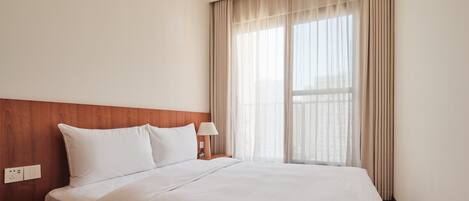 Una cama bien hecha con sábanas blancas, un cabecero de madera, una mesita de noche con una lámpara y una ventana con cortinas traslúcidas.