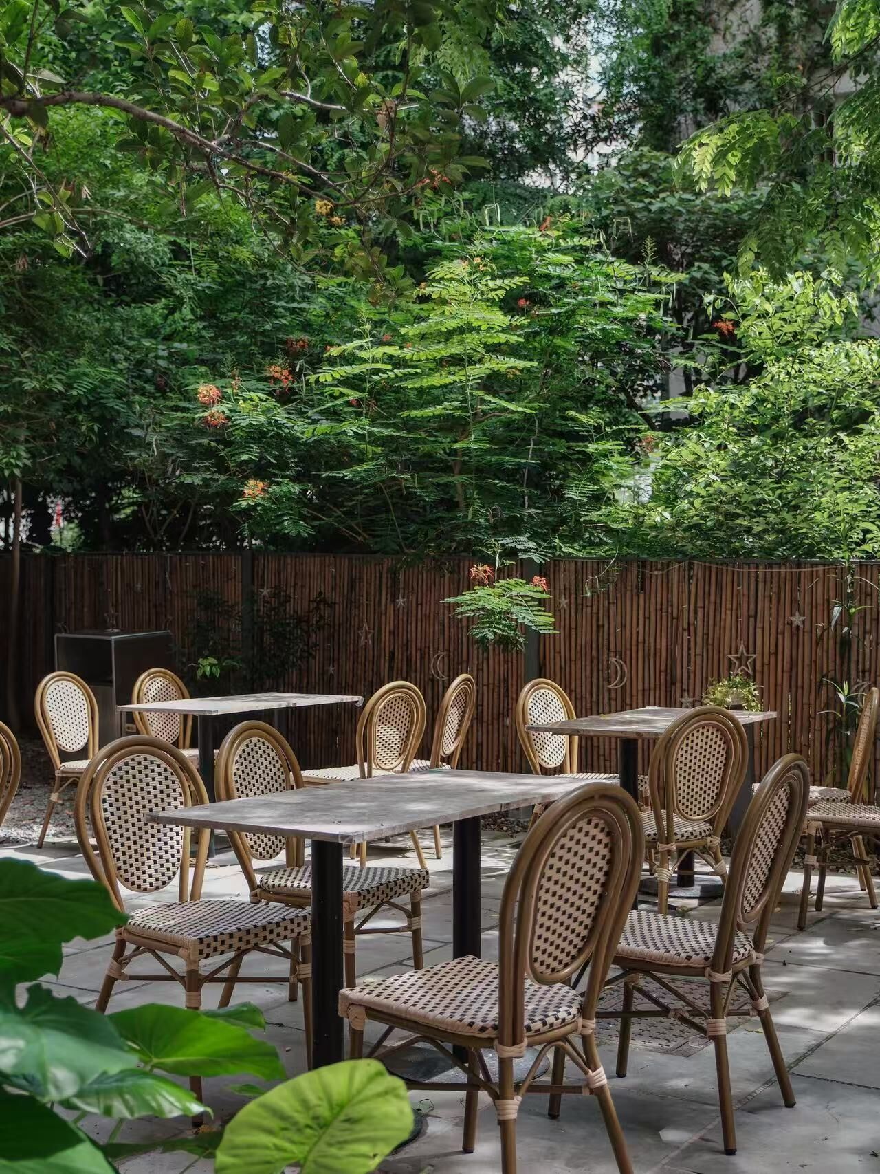 Un patio al aire libre con sillas y mesas de mimbre, rodeado de follaje denso y una valla de bambú.
