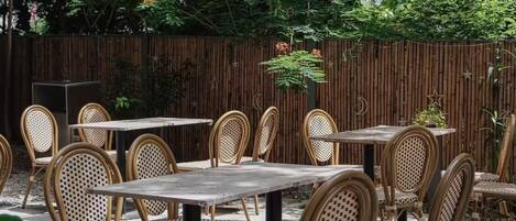 Un patio al aire libre con sillas y mesas de mimbre, rodeado de follaje denso y una valla de bambú.