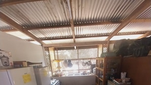 A compact kitchen with a refrigerator, stove, and sink. There is a small table with a colorful tablecloth and a chair. The kitchen has a corrugated metal roof and a window with a view outside. - Tapuhere Lodge (Huahine)
