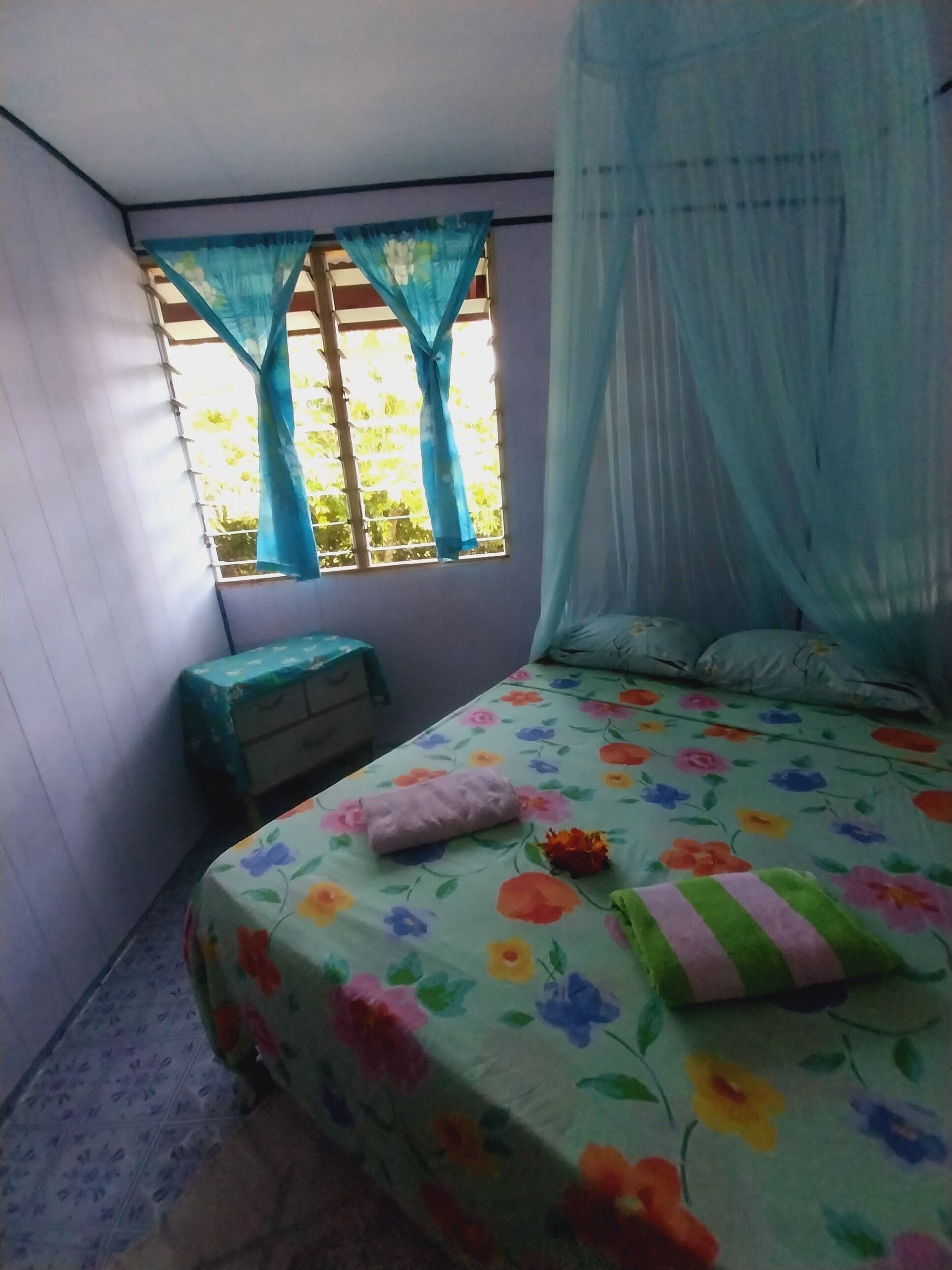 Ein Schlafzimmer mit einem Bett, das mit einem floralen Bettbezug bezogen ist, einem Nachttisch, einem kleinen Hocker und einem Fenster mit blauen Vorhängen.