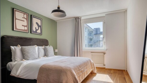 Une chambre à coucher comprenant un lit, un radiateur, une fenêtre avec des rideaux et deux œuvres d’art abstraites encadrées accrochées au mur.