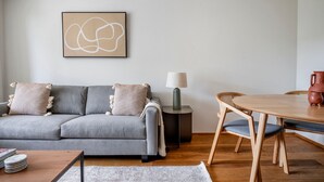 Un salon moderne avec un canapé gris, une table ronde en bois et une petite table d’appoint sur laquelle se trouve une lampe.