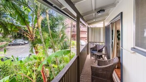 Ein Balkon mit Korbmöbeln, Blick auf eine Straße und Grünflächen, sowie ein Gebäude im Hintergrund.