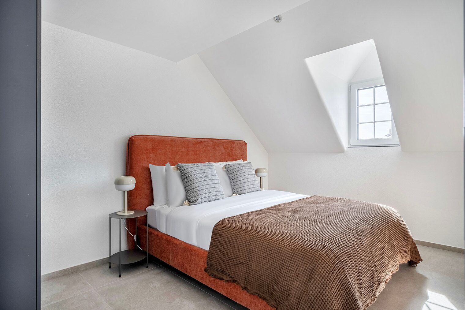 A modern bedroom with a sloped ceiling, a large window, a bed with a red headboard, and two bedside tables with lamps.
