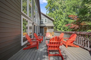 A wooden deck with red Adirondack chairs, a table, and a building with large windows. - Lazy Bear Lodge: Hot Tub, Game Room & Dogs OK (Mount Hood Village)